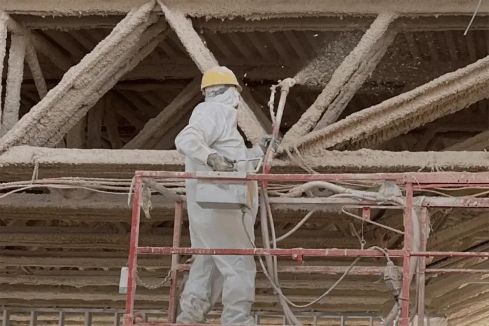 A worker spraying fireproofing onto a steel and concrete ceiling